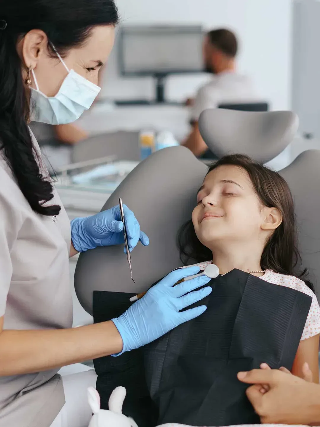 A pediatric dentist gently speaking to a young patient whose eyes are closed, relaxed during treatment.